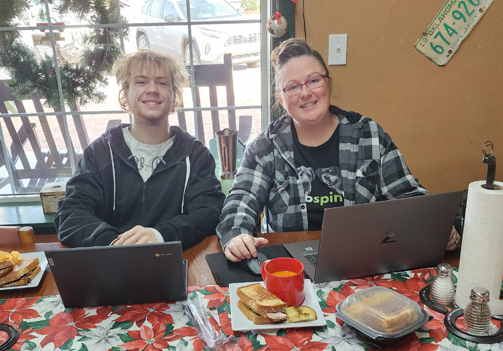 Clayton Smith and Leslie Merritt enjoying a hybrid working lunch day at The Farmer's Daughter Market. They appear to be enjoying soup and sandwiches while working on their laptops and posing for a photo.